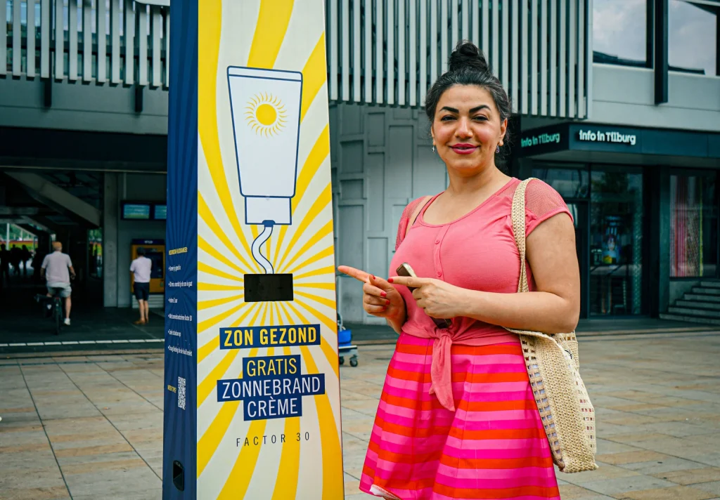 Vrouw wijst naar gele Zonnebrandpaal van Smeerkees Zonpreventie op stationsplein Tilburg
